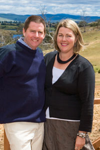 A man and a woman standing in the foreground, with a rural farm in the background.